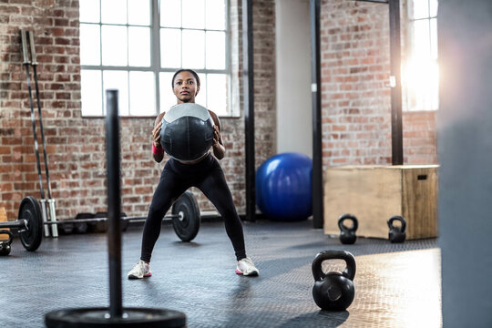 African American woman wearing sportswear holding medicine ball and squatting on rubber mat in gym