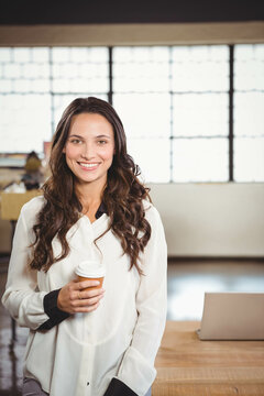 Woman standing holding coffee cup at desk wearing white blouse in open-plan office with laptop