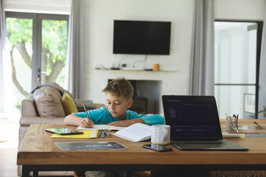 Boy writing in yellow notebook at wooden table in living room with laptop tablet and smartphone