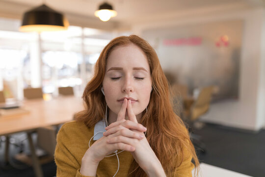 Fair-skinned woman in mustard sweater and earbuds sitting at open office desk near conference table