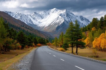 Fototapeta premium A road winds through autumn trees toward a snowcapped mountain under a cloudy sky
