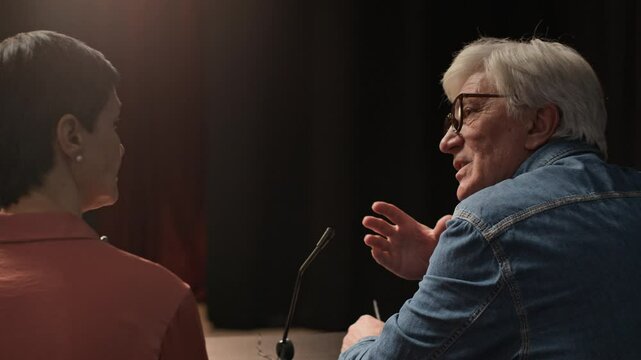 Back view of senior Caucasian man in eyeglasses sitting at table in front of stage engaged in conversation with mature woman and announcing into microphone working as judge during audition