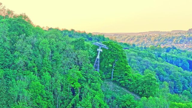 Drone captures a support tower of the Heights of Abraham cable car nestled in lush woodland near Matlock Bath, offering sweeping views of the Derwent Valley at golden hour in Derbyshire, UK.