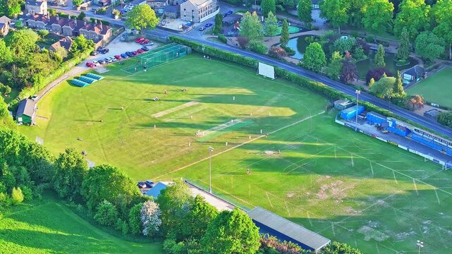 Drone shot of Matlock and Cromford Meadows Cricket Club in Derbyshire, UK, showing match in progress, lush green pitch, white-clad players, bright evening light, aerial static, wide angle