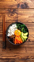 A vibrant bowl of Korean bibimbap with various colorful vegetables, rice, egg yolk, and sesame seeds is displayed on a rustic wooden table with chopsticks.