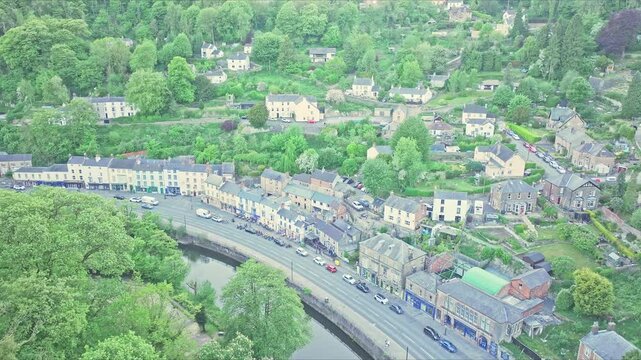 Drone shot over lively Matlock Bath, Derbyshire, UK, showing colorful buildings along the River Derwent&rsquo;s curve, surrounded by green hills and houses, vibrant spring foliage, aerial static, wide view