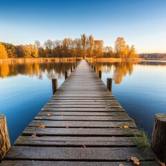 Obraz premium A long wooden pier stretches over a calm lake, surrounded by autumn trees under a clear blue sky.