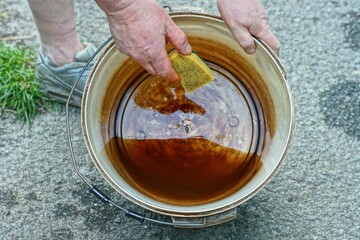 man holding dirty round industrial plastic bucket with iron handle with hands and washing with foam kitchen soft yellow sponge on asphalt in daytime on street