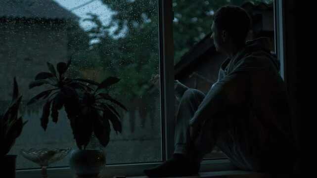 Young man sitting on window sill at rainy evening, smoking in silence. Moody contemplative atmosphere with soft warm glow.