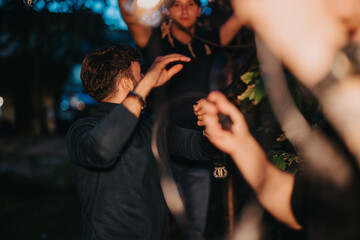 A group of friends enjoys an outdoor evening gathering. Illuminating lights create a warm and lively ambiance, surrounded by trees. The atmosphere feels joyful and celebratory in a casual setting.