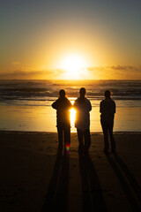 Three teenage girls silhouetted quietly on the dark sands of Muriwai Beach, gazing at a golden...