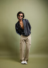 Stylish young man posing in striped shirt and cargo pants with accessories on green background.