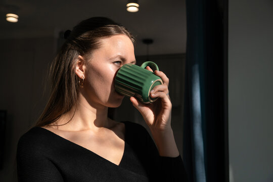 Young woman wearing a black blouse sips from a green mug, bathed in the warm glow of morning sunlight streaming through a nearby window, creating a cozy and peaceful atmosphere - Powered by Adobe