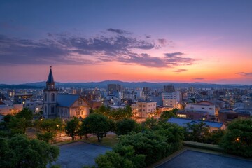 Obraz premium Cityscape at dusk with church Buildings lights mountains and pink sky fill the frame Trees and paved area are at the foreground