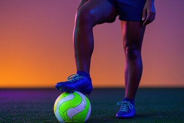 Confident female soccer player standing on green turf with foot on neon ball under vibrant colored lighting during training