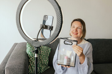 Middle-aged woman beauty blogger filming skincare product unboxing at home with ring light....