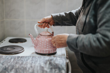 An elderly woman carefully places a pink polka-dot teapot onto an electric stove, preparing for a soothing tea moment in her warm and homey kitchen.