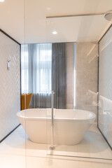 Modern bathroom featuring a white freestanding bathtub, glass partition, herringbone tile walls, a mustard yellow chair, and natural light from a large window.