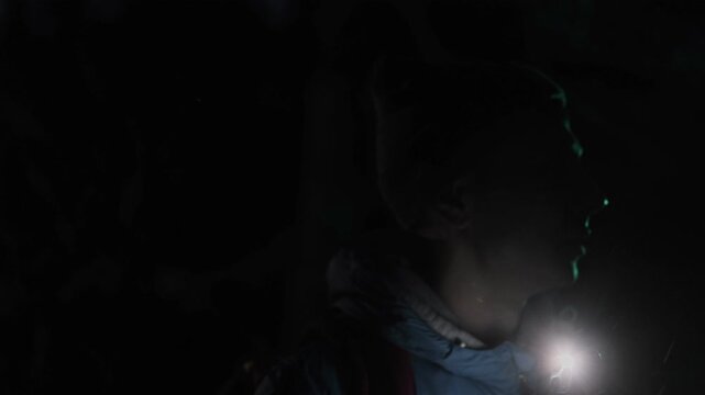 Close-up of a hiker navigating the dark forest at night, using a flashlight to find the right path under the moonlight. He scans the surroundings, trying to stay on track.