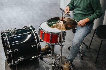 A person in a green sweater and jeans plays the drums with focus and skill indoors. The image captures the essence of music practice and dedication, highlighting the musician's relaxed style.