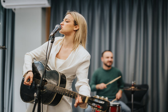 A blonde woman sings and plays the guitar while a drummer supports her in a music studio. They appear focused on creating music together during this professional band practice session.