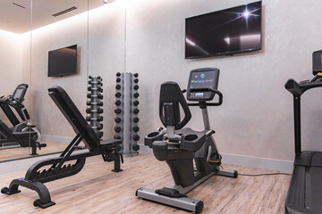 Fitness area featuring a recumbent bike, weight bench, treadmill, and dumbbell rack on wooden flooring, with mirrored walls and mounted TVs.