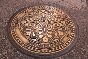 Circular manhole cover in Budapest featuring intricate floral designs, a central emblem, and metallic golden finish on textured stone pavement.