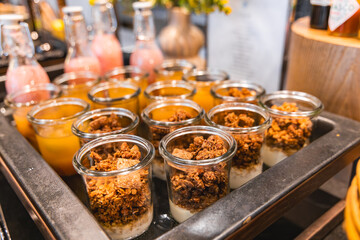 Glass jars of yogurt and granola on a tray with vibrant juice bottles in orange and pink hues, set against a blurred elegant background with flowers.