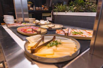 A sleek counter displays assorted cheeses, cold cuts, and garnishes with serving tongs. Warm lighting and lush greenery enhance the modern decor.
