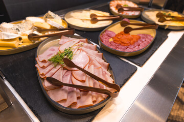 An elegant buffet setup with sliced meats, cheeses, and herbs on black slate boards. Wooden tongs and modern lighting enhance the upscale presentation.