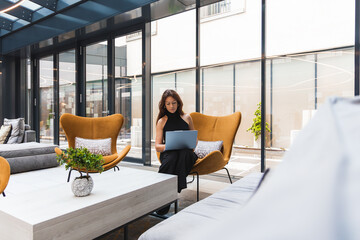 A woman sits on a mustard yellow armchair with a laptop in a bright hotel room. Floor to ceiling...