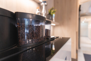 A coffee machine with transparent bean containers in a refined interior featuring light wooden paneling, minimal decor, and blurred hallway in the background.