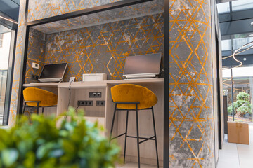 Stylish working area featuring a desk with two computers, mustard yellow bar stools, geometric wall design, natural light, and a courtyard view.