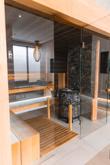 Luxurious spa interior featuring a glass walled sauna, light wood details, stone wall with central heater, tiled floor, and sleek lighting fixtures.