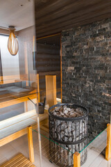 Modern sauna interior featuring sleek wooden benches, a stone wall, a central heating element with stones, and a warm wooden light fixture.