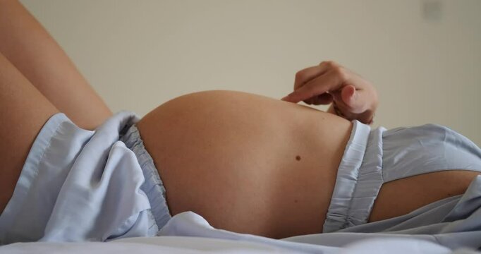 Portrait of a pregnant young woman. She lies on the bed in her pajamas and strokes her round belly in anticipation of the arrival of the baby. Third trimester of pregnancy, medical care and childbirth