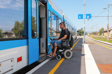 Man with disability navigates a wheelchair onto a train at a transportation hub during the day. He is focused on using a device, showcasing determination and independence.