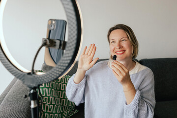Middle-aged woman beauty blogger filming skincare product unboxing at home with ring light....