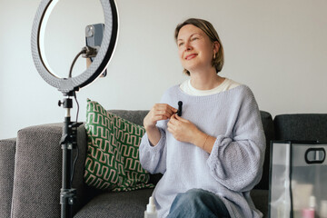 Middle-aged woman beauty blogger filming skincare product unboxing at home with ring light....