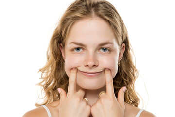 Young woman with wavy blonde hair forcing a smile by pushing her cheeks with fingers, demonstrating...
