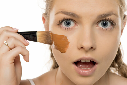 Close-up of surprised young woman applying foundation that's too dark for her skin tone, showing shocked expression while holding makeup brush against white background