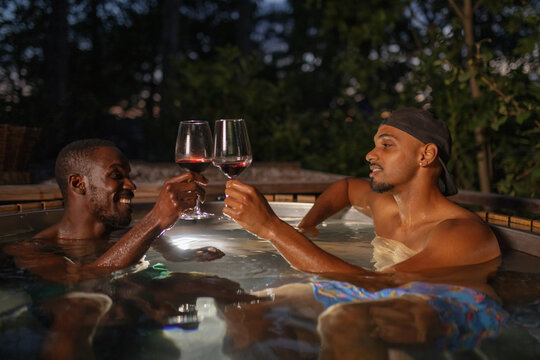 African American LGBT romantic gay couple relaxing in a private outdoor hot tub at night, enjoying glamping spa luxury drinking tasty red wine.