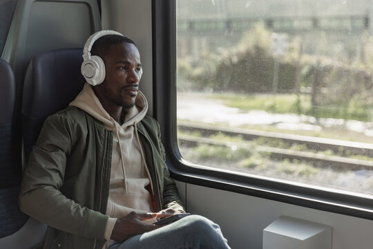 African American man sitting in the window seat on the moving train, using a smartphone and headphones to listen to music.