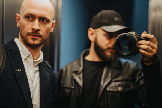 Two professional men in an office-like environment, both focusing on work while one holds a camera, portraying a collaborative and creative professional atmosphere.