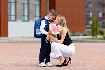 Happy mom accompanies her first-grader son to school, kissing and hugging him, back to school, the parent gives the child to the first grade. Mom meets a student from school. High quality photo