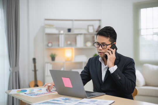 Business professional in a modern office talking on the phone while working on a laptop surrounded by charts and graphs on a desk