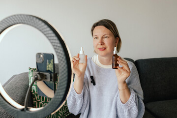 Middle-aged woman beauty blogger filming skincare product unboxing at home with ring light....
