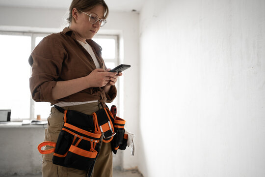 Construction worker or repairman in tool belt using smartphone in the room. profession, construction and repair