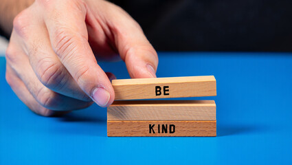 A hand places a wooden block with the word be on top of a block that says kind on a blue background
