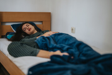 Obraz premium A young woman rests in bed, wrapped in a cozy blue blanket. The peaceful setting conveys relaxation and comfort, emphasizing a calm and serene environment.